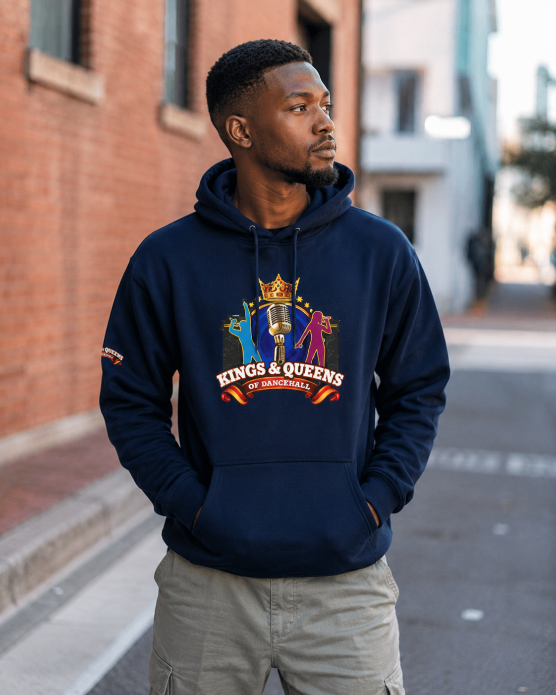 Man wearing a navy Kings & Queens of Dancehall hoodie with the crown-and-microphone crest, on a brick-lined street.