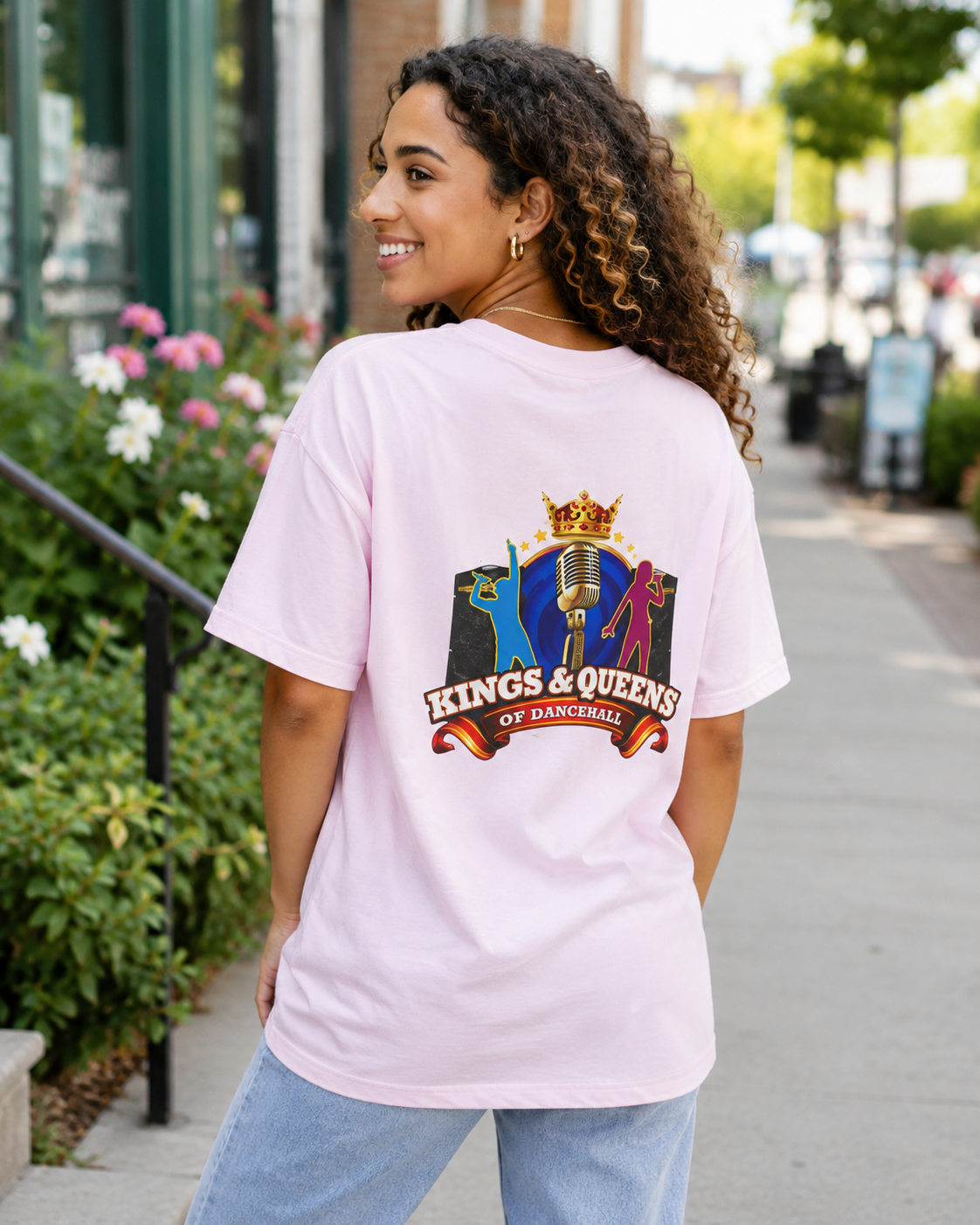 Woman seen from behind in a pink Kings & Queens of Dancehall short sleeve tee with the crest, on a sunny street.